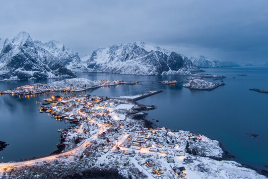 Aerial view of the snow-covered village of Lofoten with illuminated houses, winding roads, and dramatic mountains reflecting in the calm sea in Nordland, Norway.