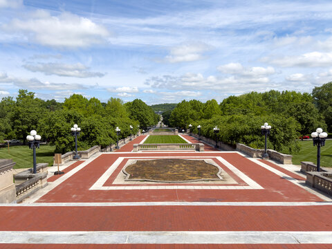 FRANKFORT, KY, MAY 6, 2026: Brick terrace with limestone borders overlooking a manicured lawn, tree-lined all&eacute;e, and globe lampposts, viewed from the Kentucky State Capitol steps under blue sky