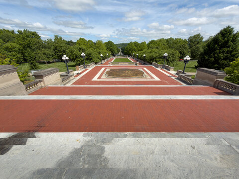 FRANKFORT, KY, MAY 6, 2026: Wide-angle view down the Capitol's marble steps and red brick terrace toward the central planting bed, lawn, and tree-lined avenue beneath scattered clouds