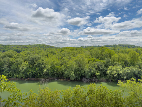 Kentucky River's green waterway winds beneath a dense canopy of leafy trees and forested hills, with a distant tower on the ridge under a sky of layered white clouds
