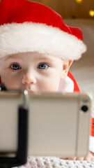 Happy baby girl in red Santa costume laughing during interactive video call. Tiny kid looking at smartphone screen with cheerful holiday child emotion. Virtual Christmas family greetings © Marina Demidiuk