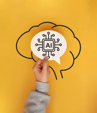 Artificial Intelligence and Human Brain Concept. Hand holding an AI symbol inside a brain outline on a yellow background.