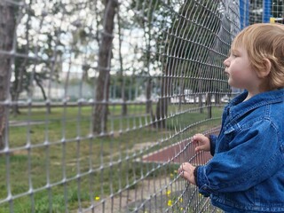 Toddler girl in denim jacket looking thoughtful near sports ground fence © Nastya