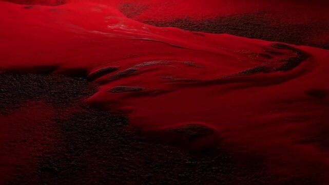 Red liquid flows and spreads smoothly across a dark textured surface with subtle reflections and soft shadows. Flowing motion creates subtle reflections and visible depth.