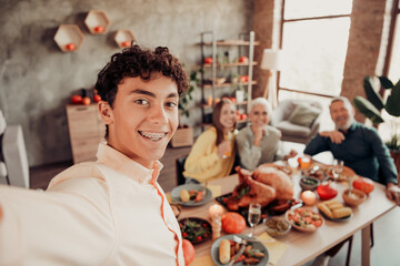 Obraz premium Teen boy taking a family Thanksgiving selfie at a holiday dinner table with relatives smiling, festive turkey, and warm home celebration