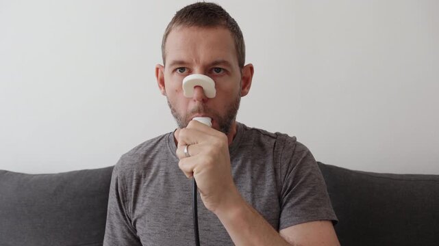 A man performs a spirometry lung test at home using a digital spirometer and a plastic nose clip on a couch
