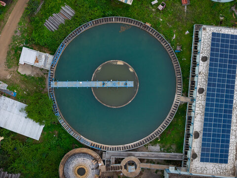 Aerial view of a circular water treatment tank with a solar panel array on the adjacent building roof surrounded by greenery in Karad, Maharashtra, India.
