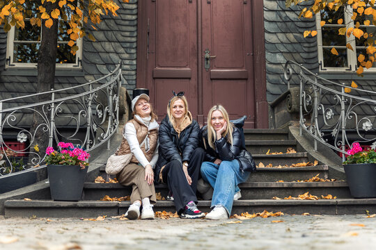Three smiling women sit on stone steps before historic doorway during autumn trip in Harz, Germany. Candid travel portrait shows friendship, tourism, leisure and warm outdoor seasonal atmosphere