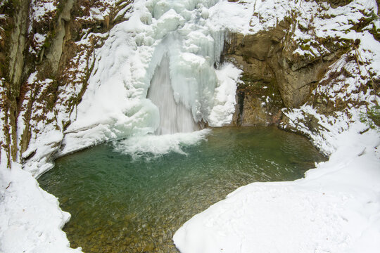 Aerial view of Casoca Pruncea Waterfall surrounded by snow-covered rocks and ice formations with a clear pool of water in Casoca, Buzau County, Romania.