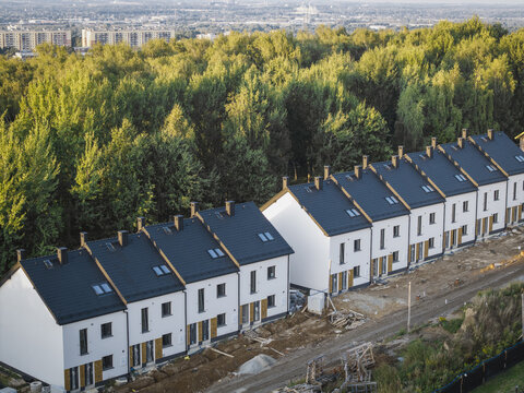 Aerial view of a row of modern white terraced houses with dark roofs under construction, situated next to a lush green forest in Wieliczka, Lesser Poland Voivode, Poland.