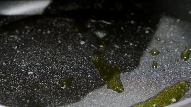 Glossy liquid swirls over speckled metallic surface with olive reflections, forming abstract wet streaks plus ripples in industrial lighting