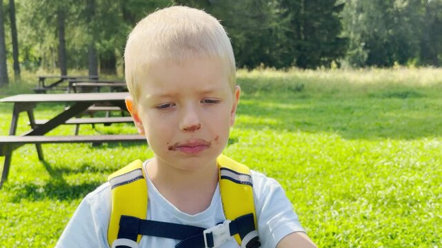 A young blond boy smiles with chocolate on his face in a green park. He feels happy, playful, and joyful during this fun outdoor snack break.