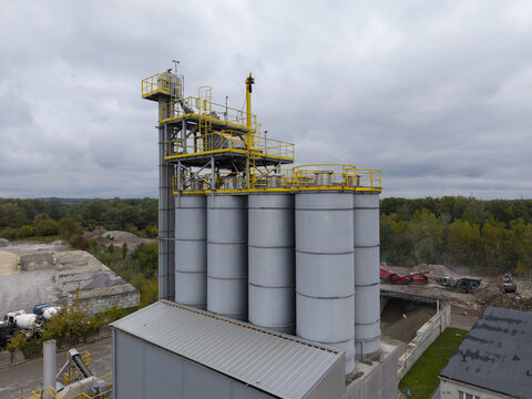 Aerial view of the asphalt plant with tall grey storage silos and yellow metal platforms surrounded by gravel piles and industrial equipment in Krakow, Lesser Poland Voivodeship, Poland.