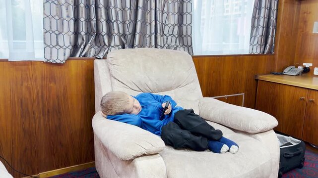 A relaxed young boy rests comfortably in a soft armchair holding a remote control. He enjoys a cozy, calm evening inside a cruise ship cabin docked in Saint Petersburg.