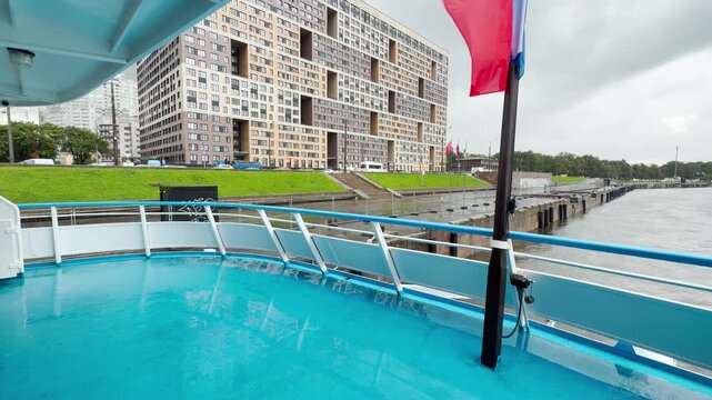 A cruise ship glides along the Neva River in Saint Petersburg. The blue deck offers a peaceful view under a calm, gloomy sky.