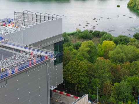 Aerial view of a modern data center building featuring rooftop scaffolding and metallic cladding, situated next to a tranquil lake and lush green woodland in the United Kingdom.