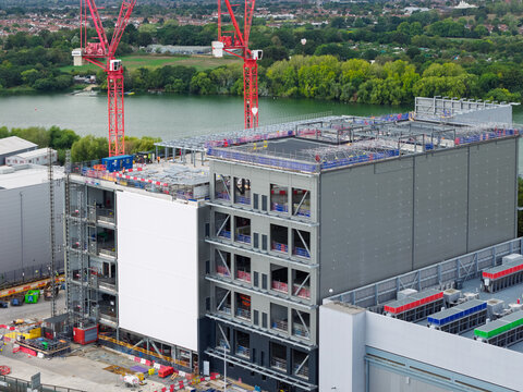 Aerial view of a large data center under construction with steel framework and red cranes situated near a lake in the United Kingdom.