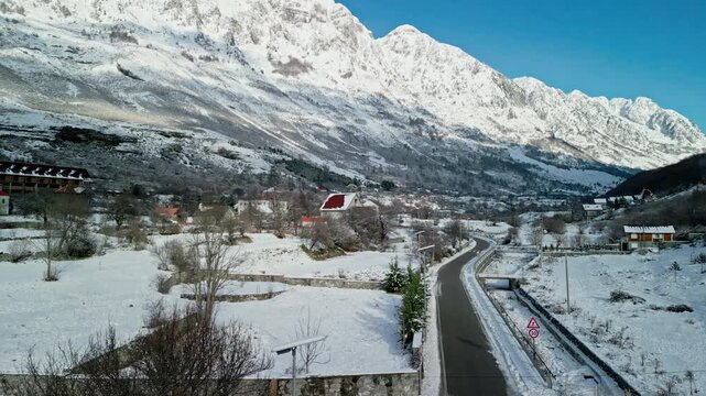 Albania, Boge 11 01 2026: Drone video ascending over snow-covered village houses and guesthouses along the frozen Prroni i Thate riverbed with snowy Albanian Alps in background.