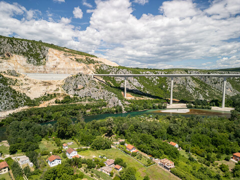 Aerial view of Pocitelj Bridge spanning the Neretva River, surrounded by green hills, rocky terrain, and houses with red roofs in Pocitelj, Federation of Bosnia, Bosnia and Herzegovina.