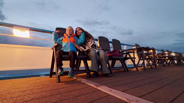 A mother and her happy son sit together on a cruise ship deck. The boy playfully flexes his arms as they sail along the Neva River at twilight. They feel joyful, cozy, and peaceful.