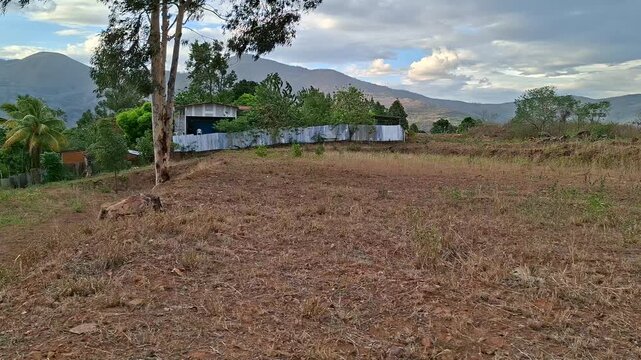 Scenic view of a rustic countryside house with a corrugated metal fence, surrounded by green trees, eucalyptus, and a dry open field in the foreground. In the background, majestic green mountains stan