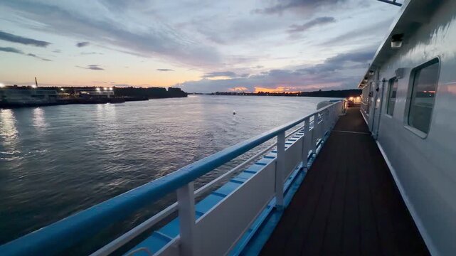 A cruise ship deck glides smoothly along the wide river. The glowing sunset sky reflects off the calm water. The scenic evening view creates a peaceful and nostalgic feeling.
