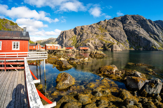 Traditional red rorbuer stand along the rocky coast of Nusfjord, surrounded by clear water and massive rocky mountains.