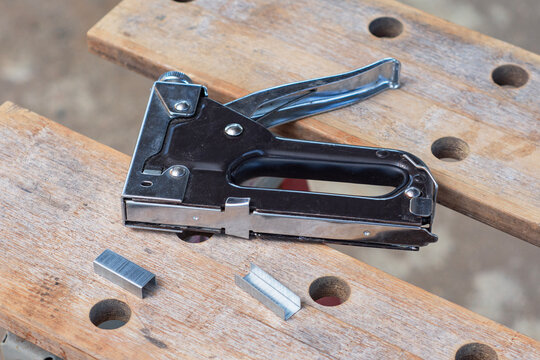 Heavy duty steel staple gun with metal staples on wooden workbench. Furniture upholstery, carpentry fastening, DIY home repair, construction tacker tool and woodworking concept.