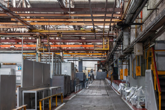 Wide-angle view of industrial factory interior with long corridor of metal beams, pipes, cables and machinery in orange, yellow and gray tones. Industrial infrastructure and manufacturing environment.