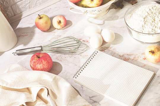 A bowl of apples, on the kitchen table, with baking ingredients, and a notebook, morning, no people,