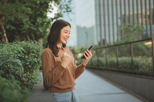 Excited young woman celebrating success while looking at smartphone outdoors, smiling with joyful fist gesture after positive news, achievement