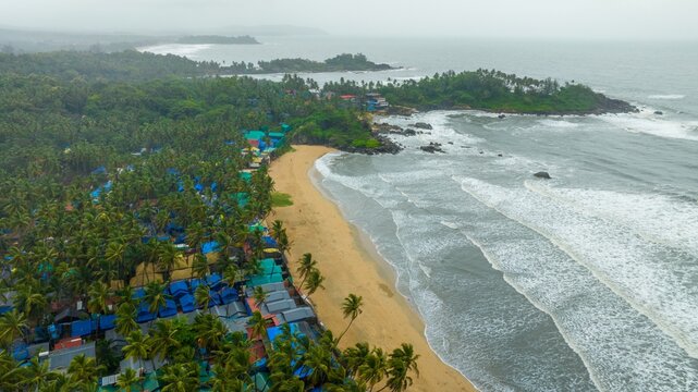 Aerial view of Palolem Beach with its golden sands, dense palm groves, and rows of blue-roofed beach huts along the coastline in Canacona, Goa, India.