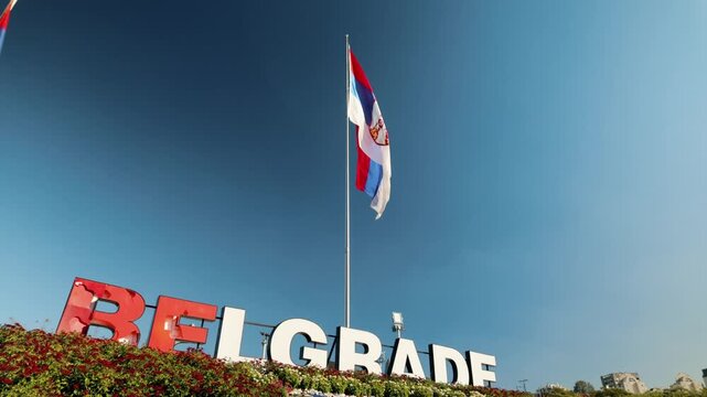 Large Belgrade city sign stands beneath a Serbian flag against a clear blue sky. Urban landmark represents tourism, travel, identity and city promotion.