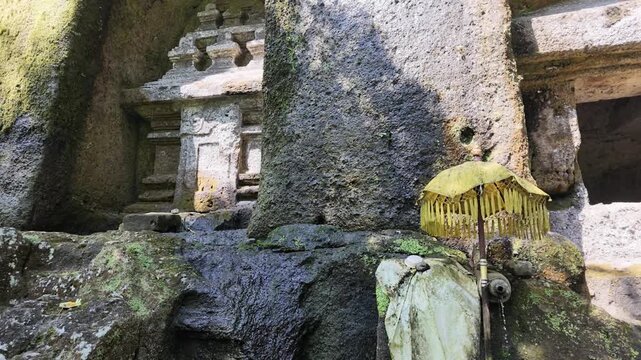 Ancient rock carved shrine at Gunung Kawi temple in Bali featuring flowing spring water and a traditional yellow ritual umbrella, cultural heritage and sacred spiritual site for travel documentary