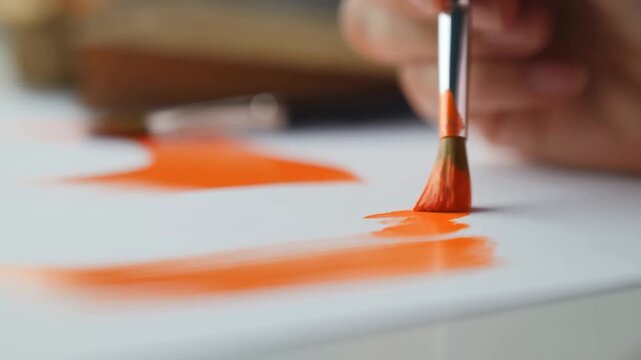 Artist hand applies bright orange paint with a brush to white paper in a close-up studio scene. Art education, painting tutorials, and creative workshop promotion.