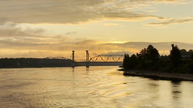 A bridge spans across the wide river during sunset. A peaceful and serene feeling fills the warm evening environment.