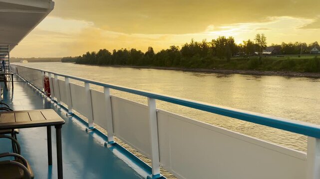 A cruise ship glides along a quiet river past lush trees during a golden sunset. The warm evening sky creates a serene view. Travelers feel peaceful and content in this calm setting.