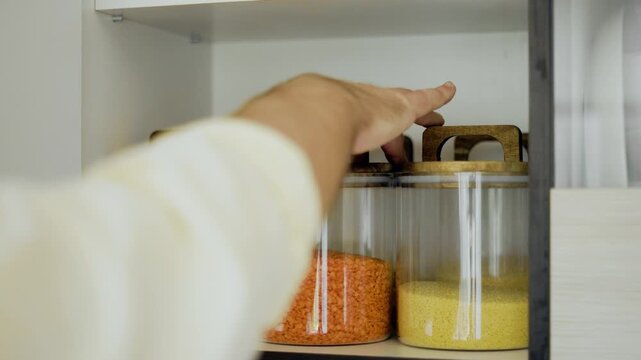 man opens a kitchen cabinet and takes a glass jar with red lentils.Organized pantry food storage with grains,rice,cereals,couscous,and clean ingredients for healthy cooking.High quality 4k footage