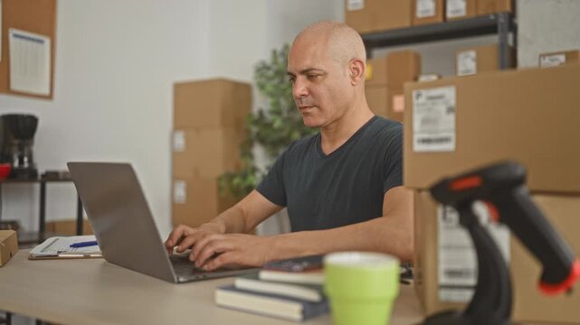 Man holding smartphone to ear and typing on laptop amid stacked packages and barcode scanner on a table in a building; focused productivity.