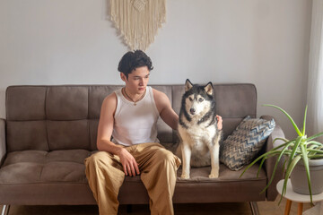 Teenage boy sits on sofa beside siberian husky in bright living room, friendship needing no words at all, friendship without any condition. Wide shot. Concept of friendship of teenager and his dog © iwavephoto