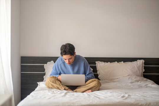 Teenager leans forward reading from laptop screen on soft bedding, student whose classroom became screen, hour that never feels short. Wide shot. Concept of student working from home bedroom