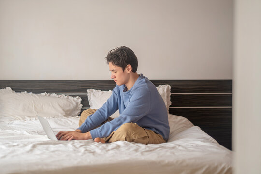 Teenage boy leans low over laptop typing homework on white bed, homework done without any help, honest look at distance learning. Wide shot. Concept of homework done from own bedroom