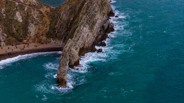 Aerial view of Durdle Door showing the limestone arch, turquoise sea waves, and a pebble beach with people walking along the coast in Wareham, England, United Kingdom.