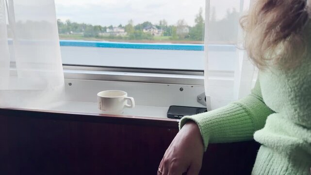 A woman rests by a cruise ship window facing a calm river. A warm cup sits beside her during the quiet trip. The peaceful view fills her with cozy relaxation and joy.