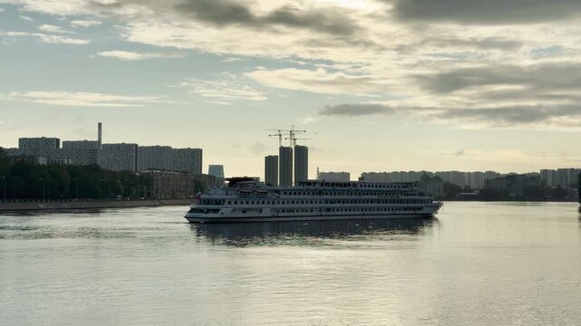 A cruise ship sails gracefully down the Neva River past the skyline of Saint Petersburg. The quiet morning water reflects soft light under a cloudy sky. This serene journey brings feelings of peace