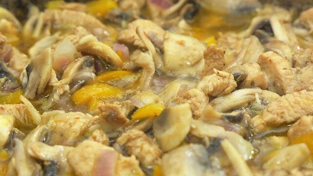 In a deep cast-iron skillet, onions simmer with chicken in a sauce and spices, along with mushrooms, and are stirred with a spoon. Medium shot.