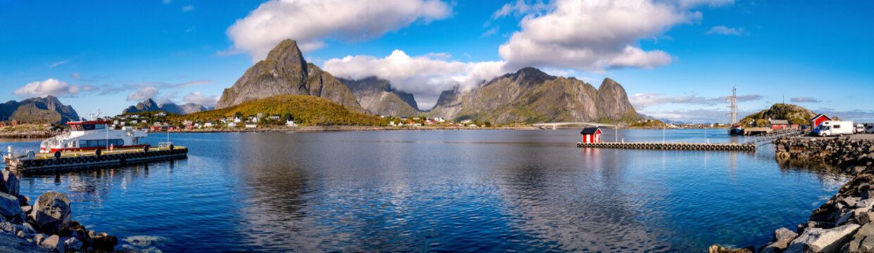 Wide panoramic view over Reine with harbor, sea, houses and distinctive mountains beneath blue sky in the Lofoten Islands.