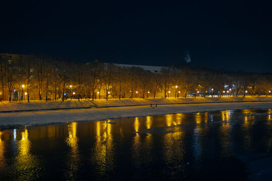 Winter night landscape of the Uzh River in Uzhhorod with warm city lights reflected in the water.