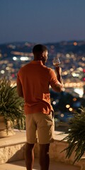 Fototapeta premium Man holding a wine glass on a terrace with blurred city lights at dusk for poster background and lifestyle or travel banner design