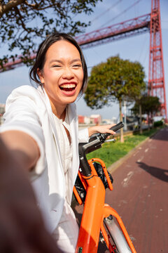 Smiling woman take bicycle selfie beside suspension bridge